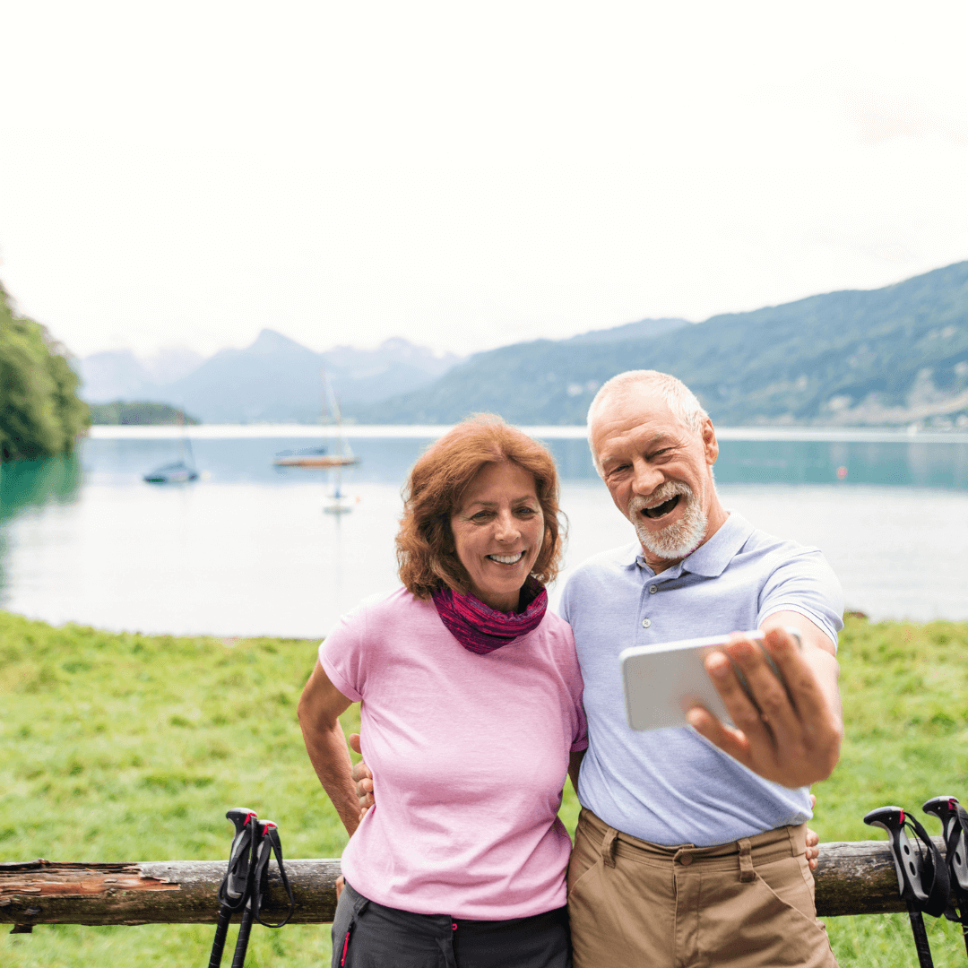 Older couple taking a selfie by a serene lake with mountains in the background, smiling and enjoying the scenery. - Home Instead