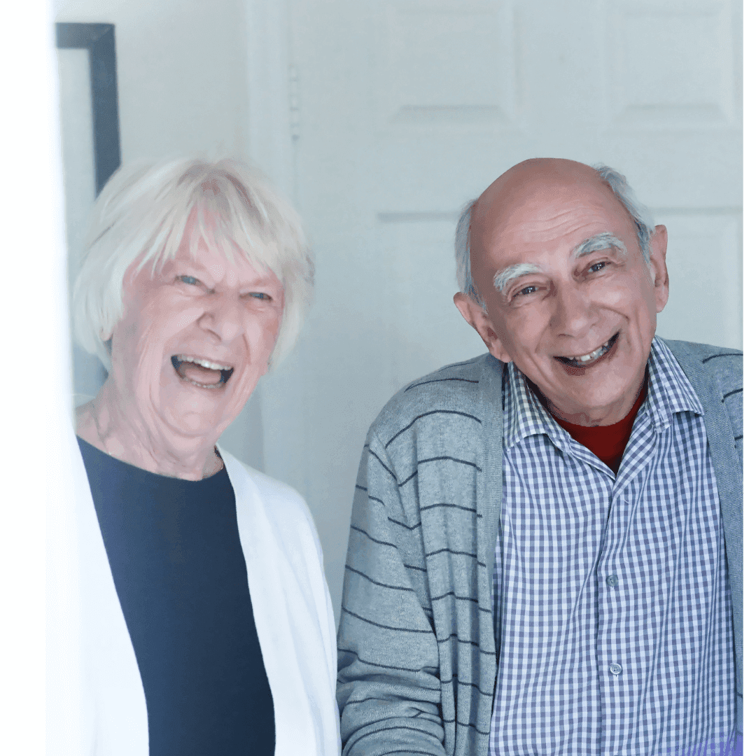 An elderly woman and man smiling and standing indoors, the man in a striped cardigan, and the woman in a white sweater. - Home Instead