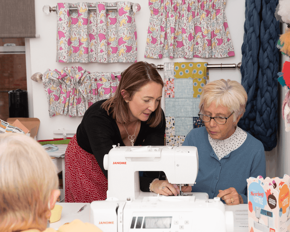A woman helps an older woman use a sewing machine in a colorful craft room with patterned curtains. - Home Instead
