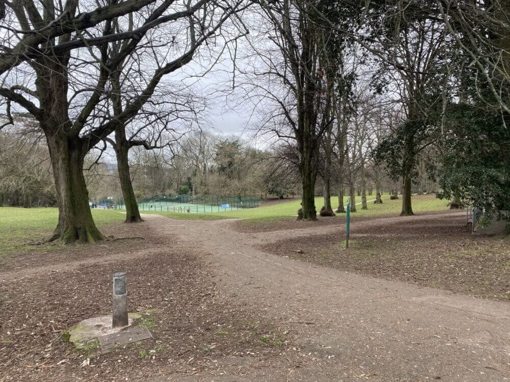 A path in a park with bare trees, leading to a fenced tennis court in the distance under a cloudy sky. - Home Instead