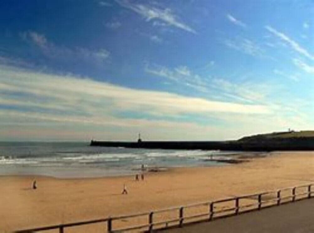 A sandy beach with a few people, a railing, and a distant pier under a blue sky with wispy clouds. - Home Instead