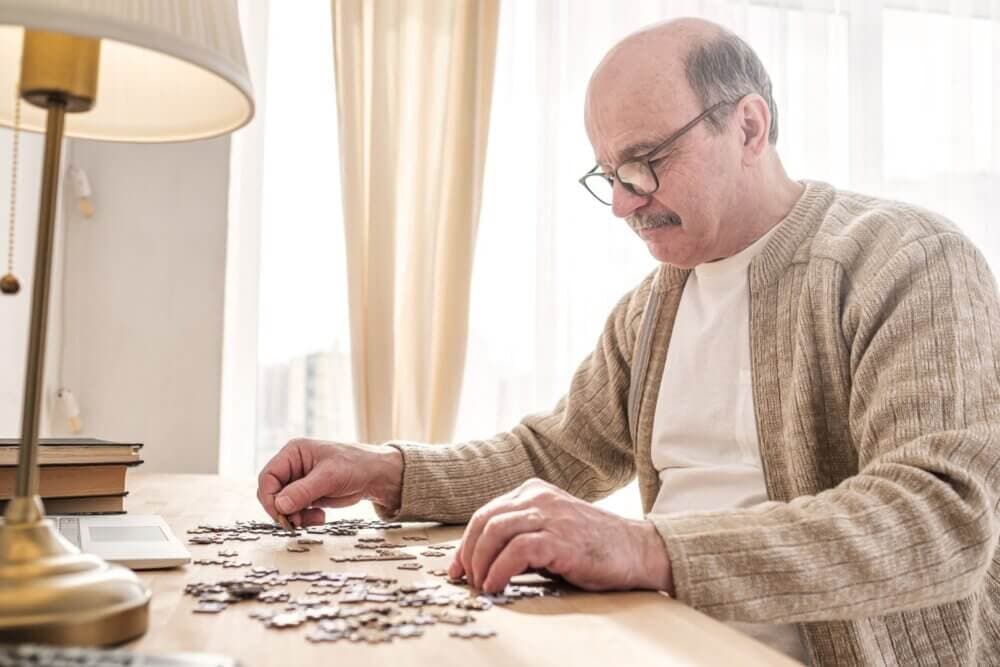 An elderly man wearing glasses and a beige cardigan works on a jigsaw puzzle at a wooden table by a window. - Home Instead
