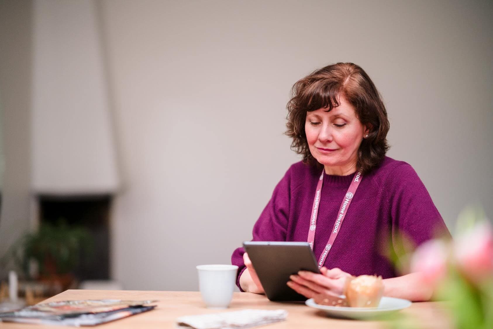 A woman in a purple sweater reads on a tablet at a table with a cup, magazine, and plate with a pastry. - Home Instead
