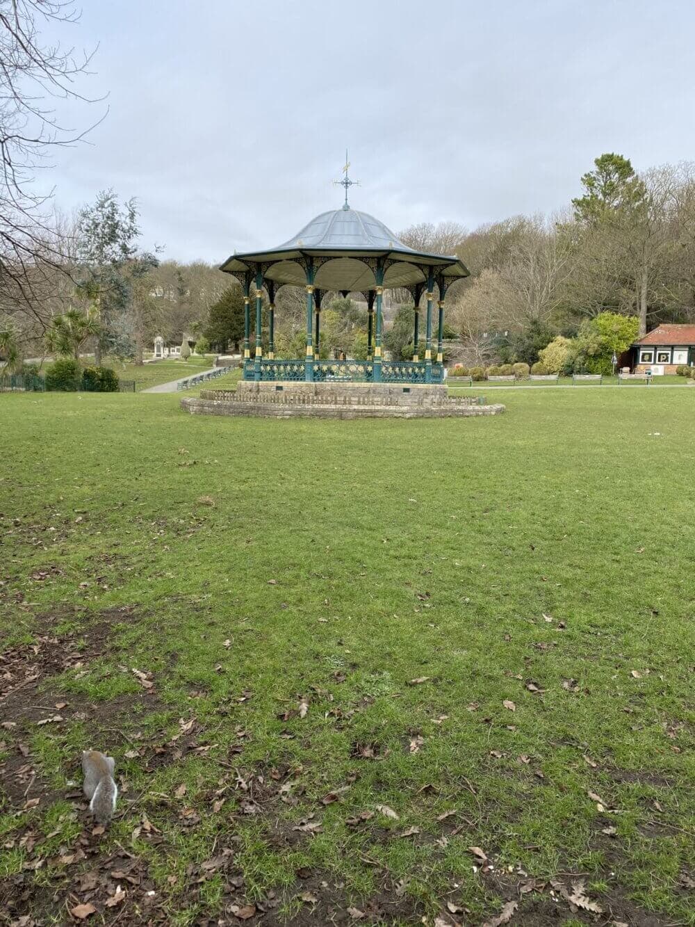 A green park with a pavilion in the center, surrounded by trees and a squirrel in the foreground. - Home Instead