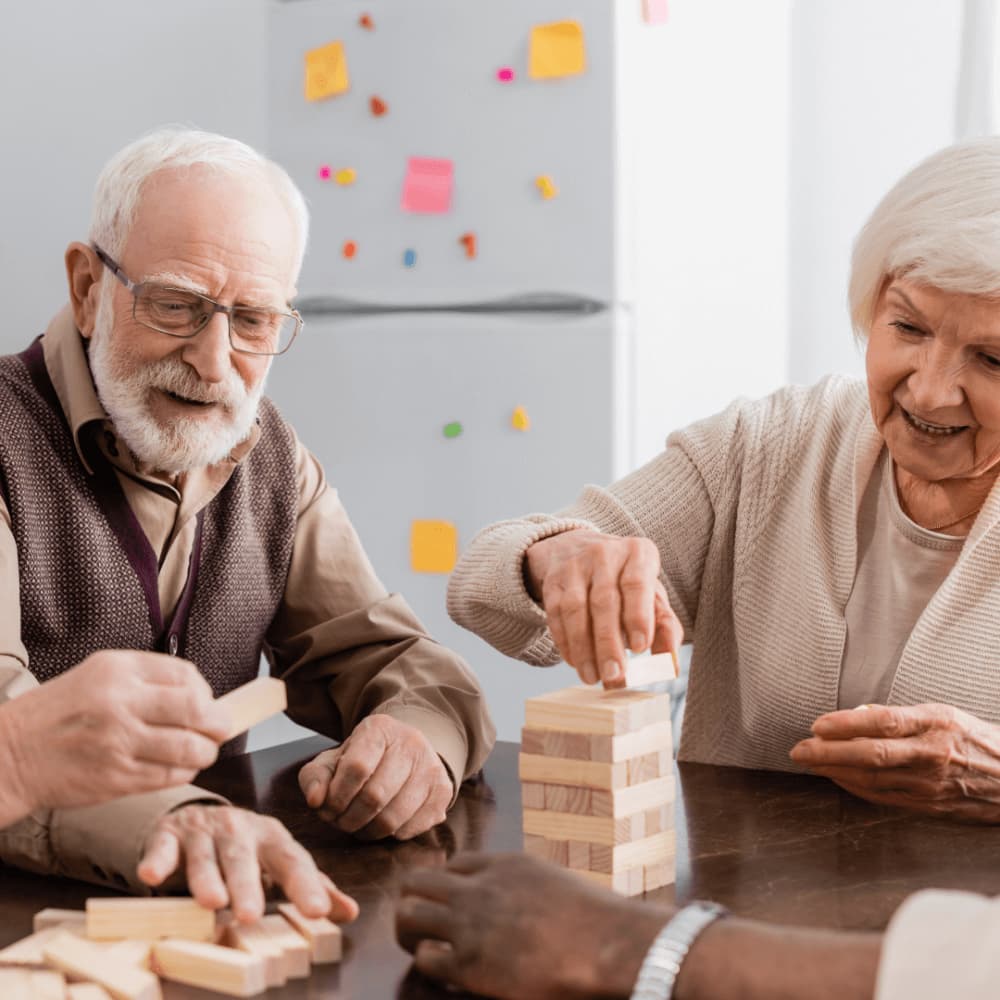 Two elderly people playfully stacking wooden blocks at a table, with a fridge covered in colorful notes in the background. - Home Instead