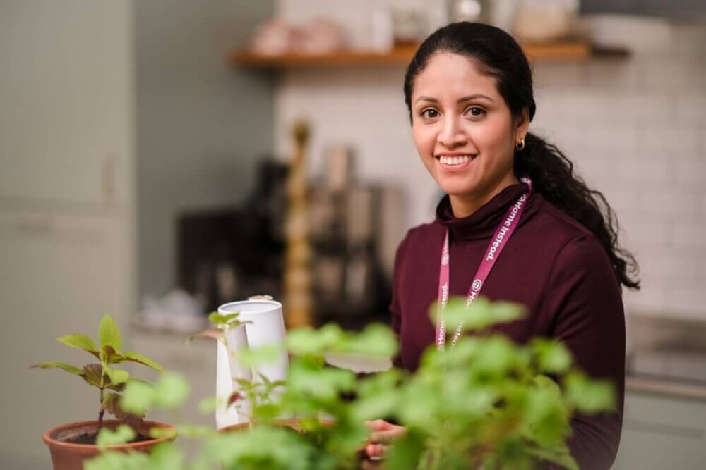 A woman with a lanyard smiles while standing in a kitchen near some potted plants. - Home Instead