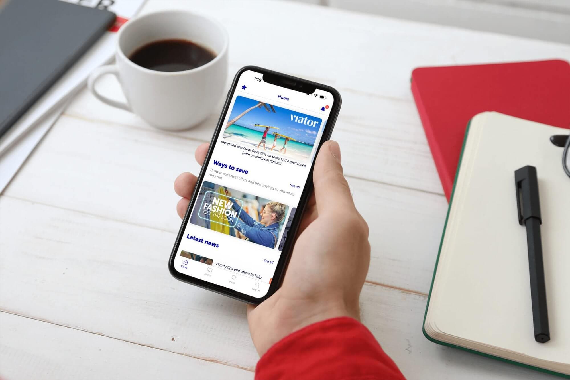 Person holding a smartphone displaying a travel booking app; desk with coffee, notebook, and pen in the background. - Home Instead