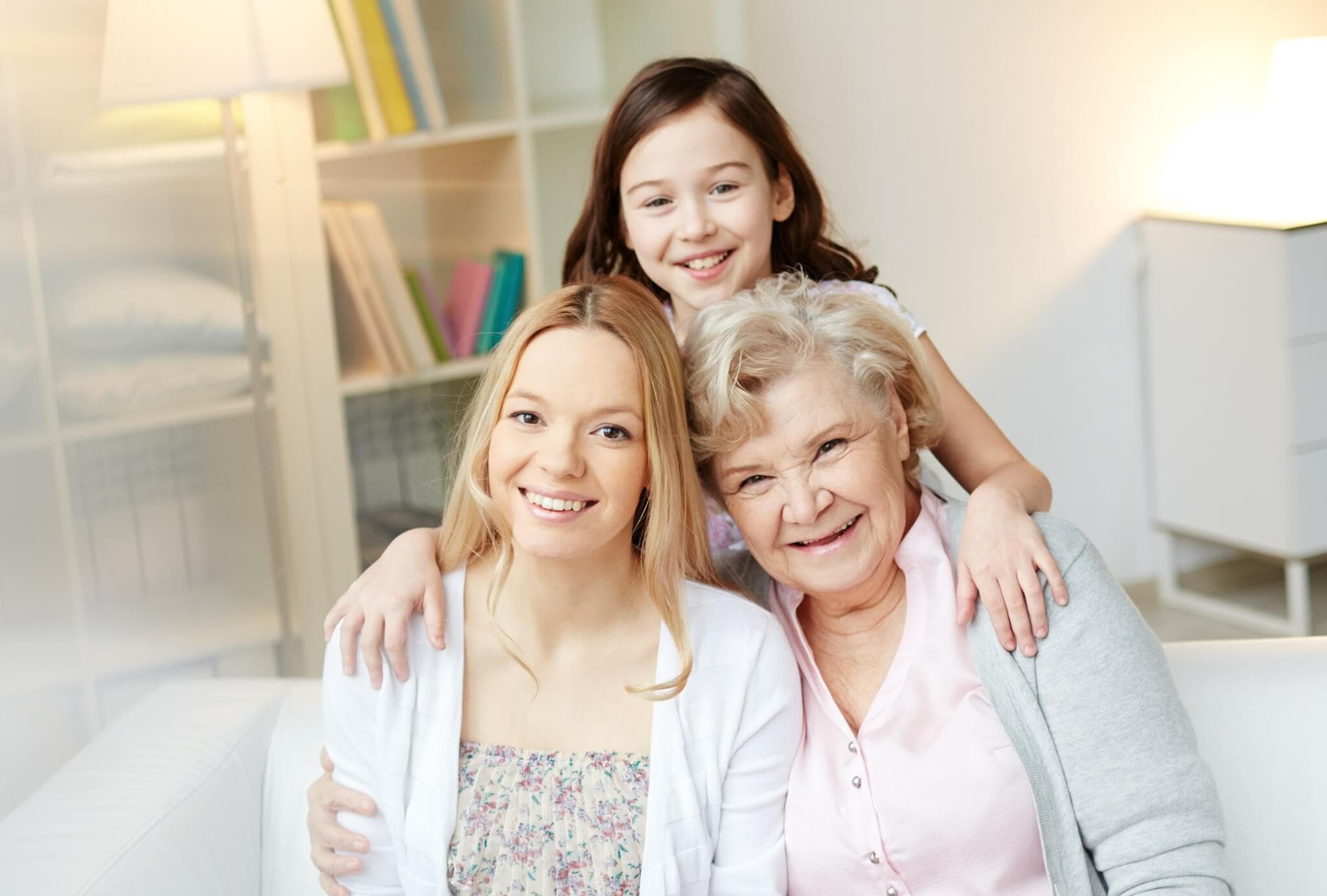 Three generations: a smiling young girl stands behind her seated mother and grandmother, all posing warmly in a cozy living room. - Home Instead