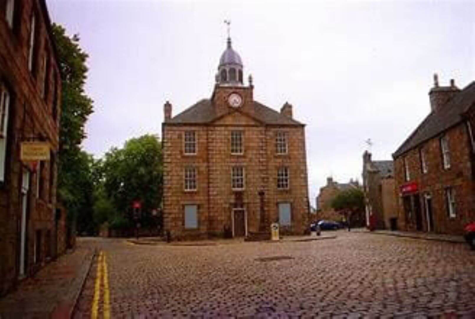 A historic, multi-story brick building with a clock tower sits at a cobblestone street intersection in a quaint town. - Home Instead