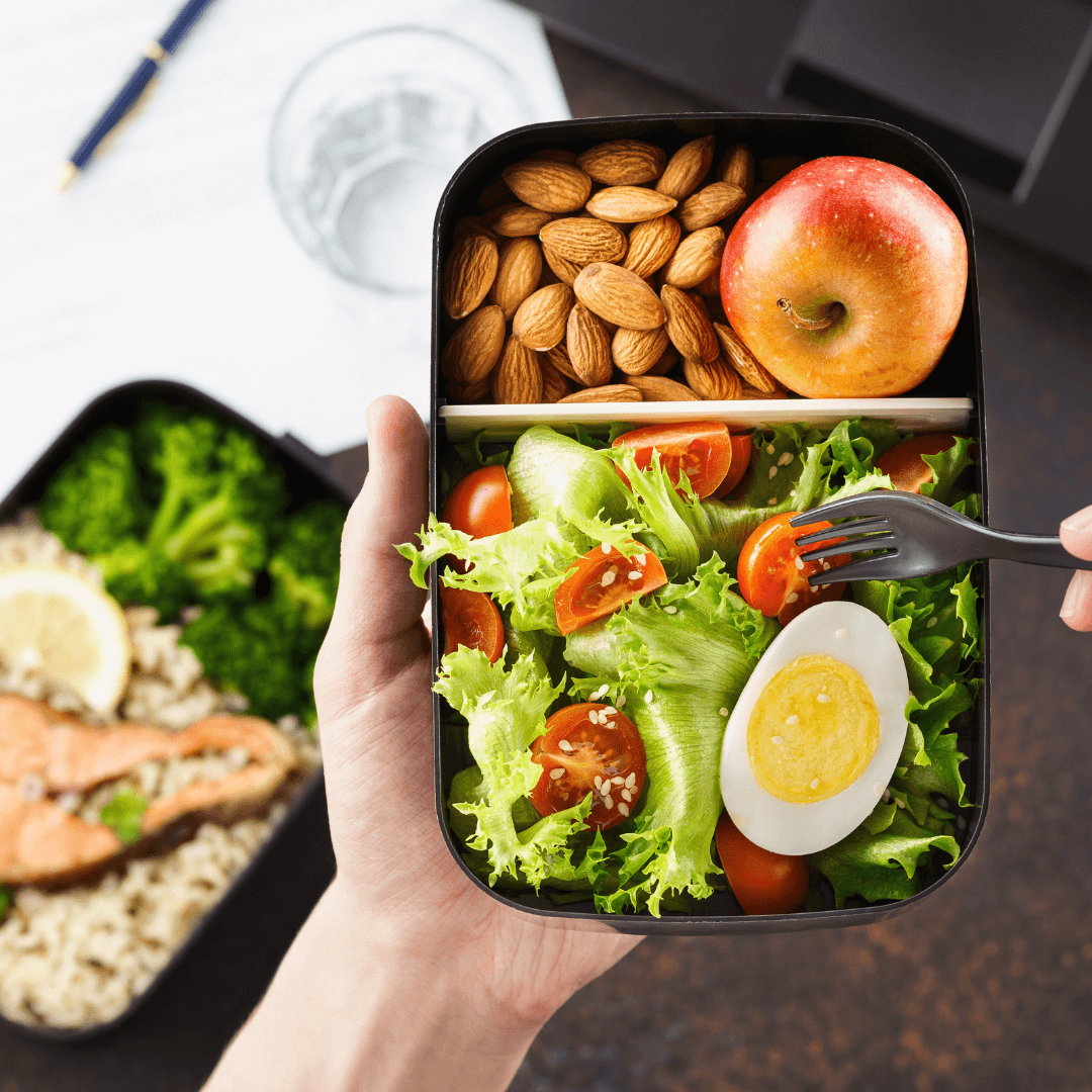 Hand holding a bento box containing a salad, hard-boiled egg, almonds, and an apple with a meal in the background. - Home Instead