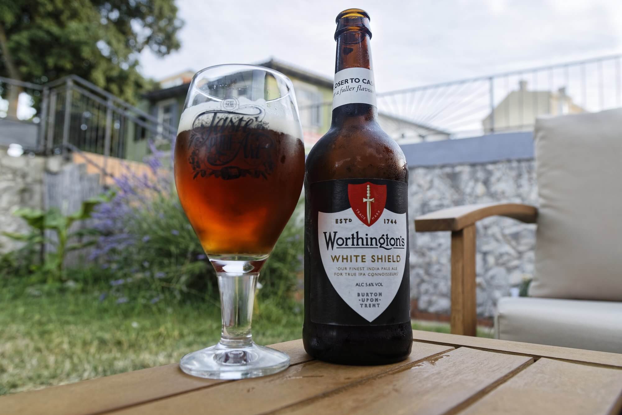 A Worthington's White Shield beer bottle sits on a table beside a filled beer glass in an outdoor setting. - Home Instead