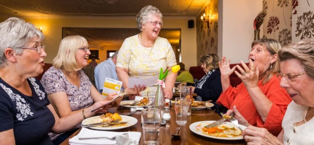 A group of older adults enjoys a meal together, laughing and engaging in lively conversation around a dinner table. - Home Instead