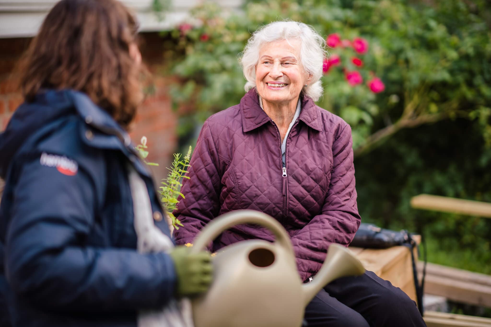 Elderly woman smiling at another person holding a watering can in a garden setting with flowers in the background. - Home Instead