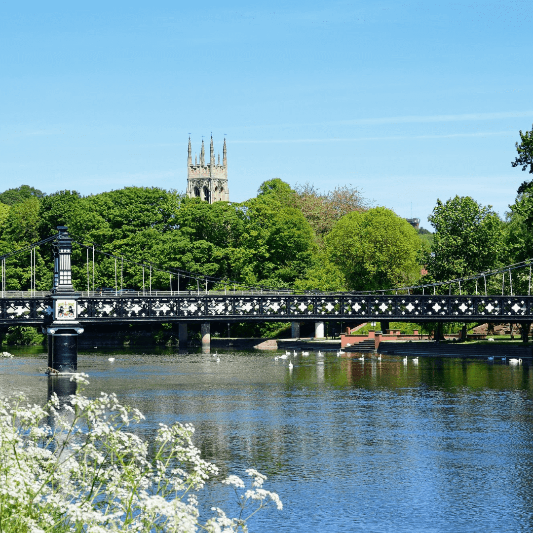 A black metal bridge spans a calm river, with a church tower rising above the lush green trees in the background. - Home Instead