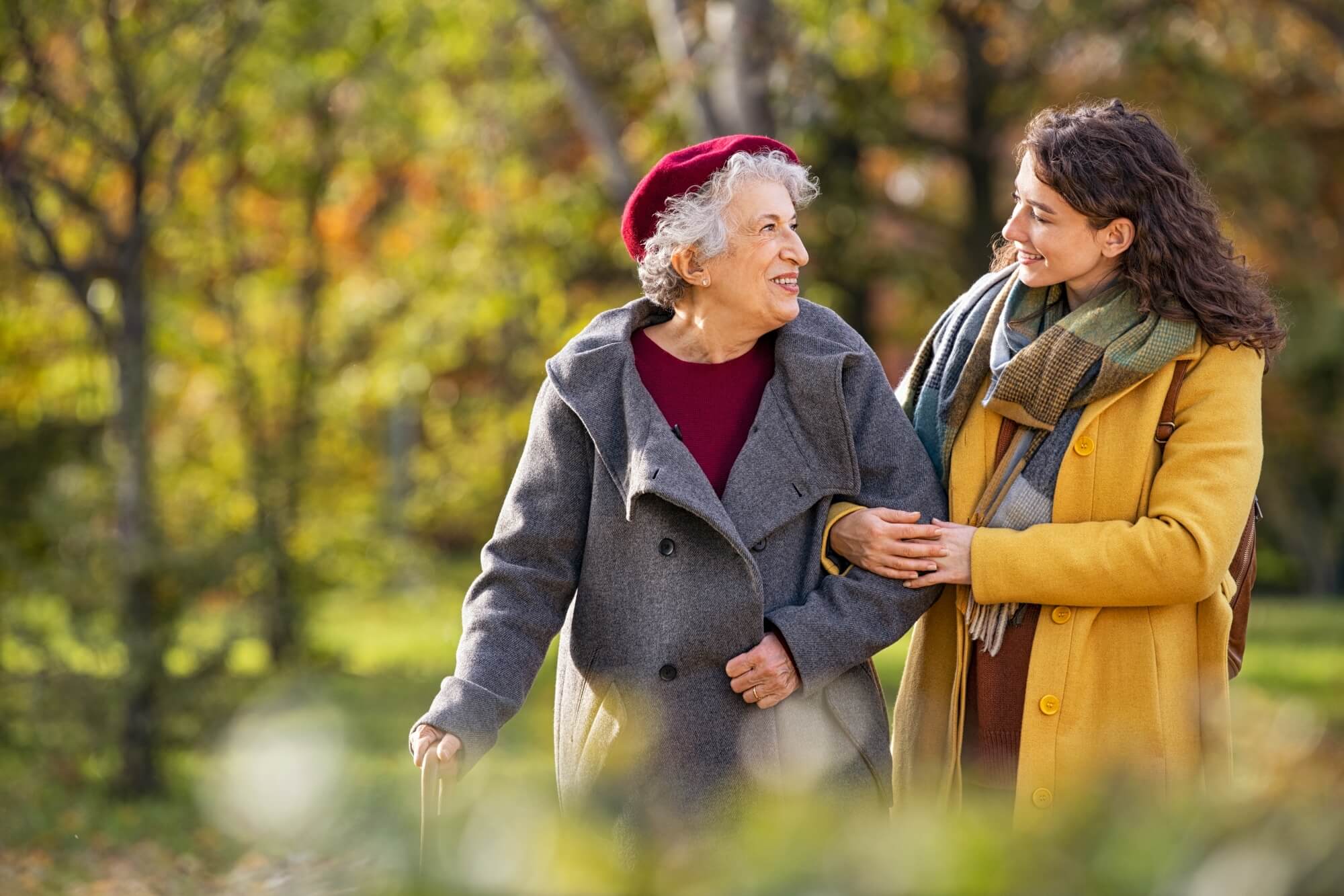 Elderly woman with a cane and young woman walk arm-in-arm, smiling, in a park with autumn foliage. - Home Instead