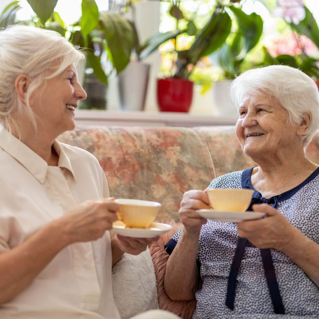 Two elderly women smiling and enjoying tea together on a floral couch with green plants in the background. - Home Instead