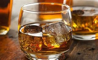 A glass of whiskey with ice cubes on a wooden surface, with another glass in the background. - Home Instead