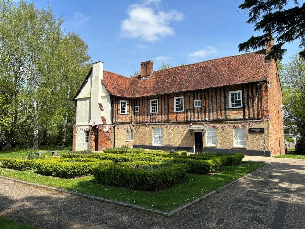 A two-story historic building with a red-tiled roof, surrounded by a trimmed garden and trees on a sunny day. - Home Instead