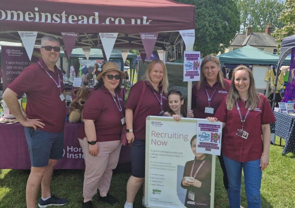 A group of people in maroon shirts poses at a recruitment booth outdoors under a tent with "Home Instead" signage. - Home Instead