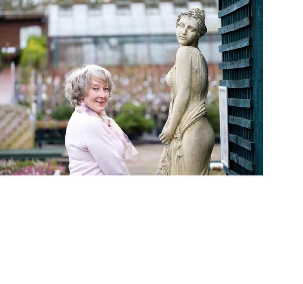 Elderly woman smiling and posing beside a classical-style statue in an outdoor garden setting. - Home Instead