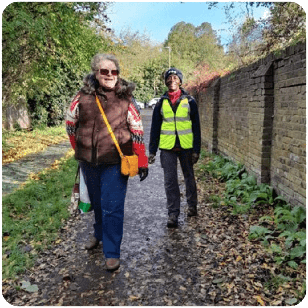 Two elderly women smiling and walking on a leaf-strewn path, one in a reflective vest, next to a brick wall and trees. - Home Instead