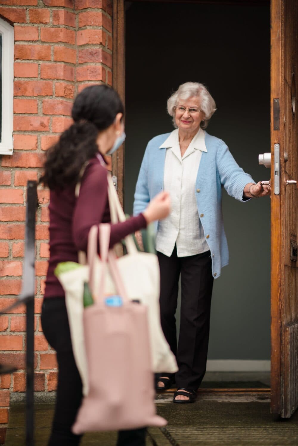 An older woman holds the door open for a younger person carrying bags, standing outside a brick building. - Home Instead Bournemouth & Christchurch