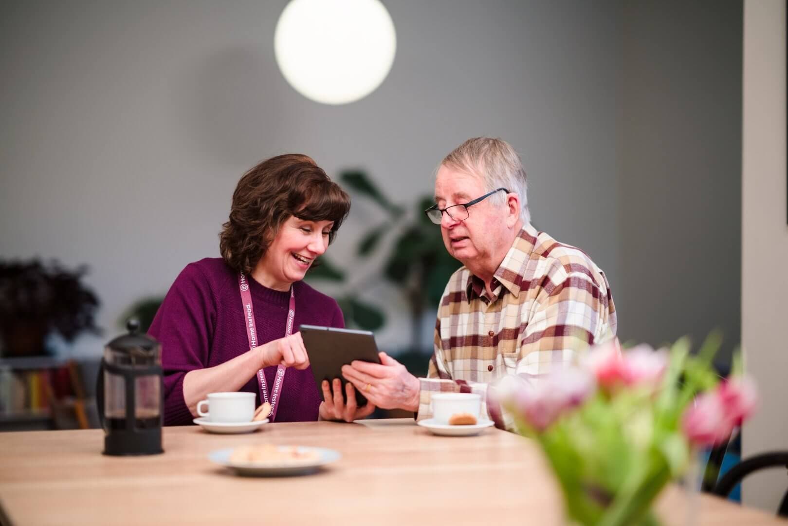 A woman and an elderly man sit at a table, smiling and looking at a tablet together. - Home Instead