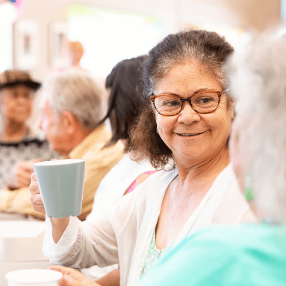 Elderly woman with glasses smiling and holding a mug, sitting among a group of seniors. - Home Instead