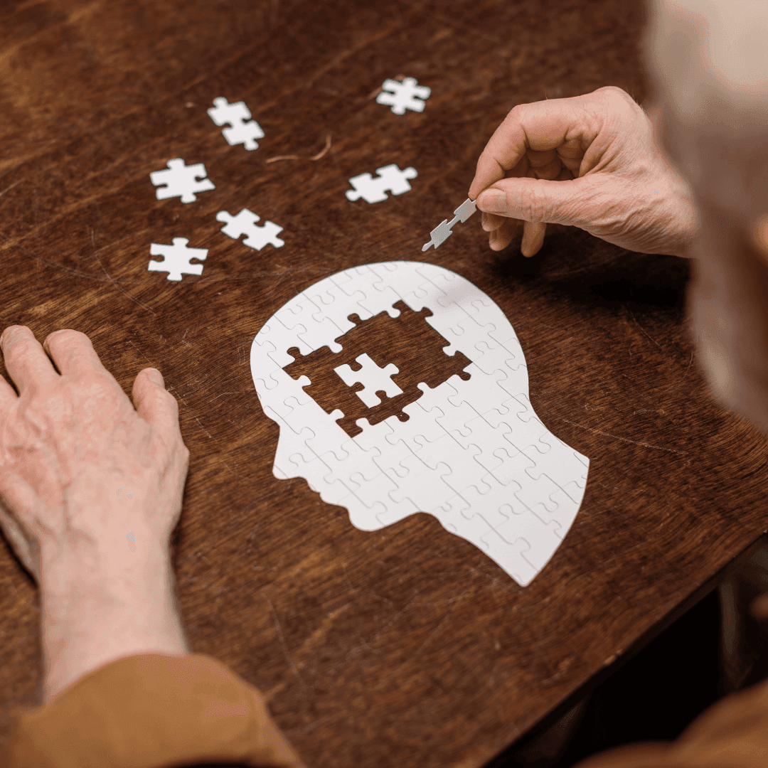 A person assembling scattered puzzle pieces into a brain-shaped puzzle on a wooden table, implying mental health. - Home Instead