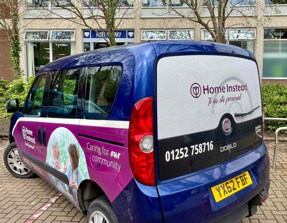 Purple and blue "Home Instead" van with contact number, parked outside a building with trees and a pharmacy sign. - Home Instead