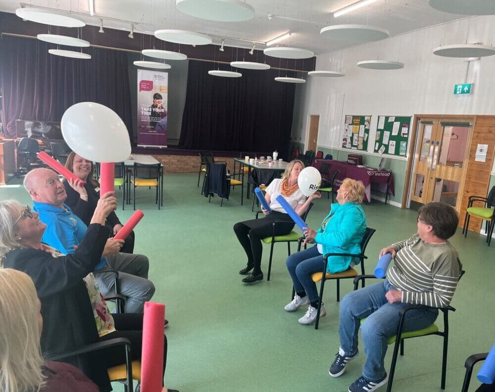 A group of people sitting in chairs, playing with foam noodles and balloons in a well-lit room. - Home Instead