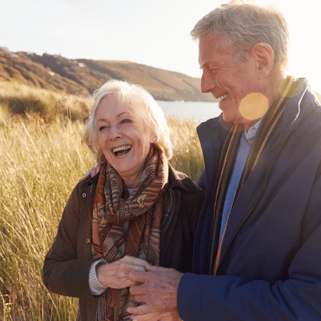 Older couple laughing and holding hands while standing in a grassy field by the water, with hills in the background. - Home Instead