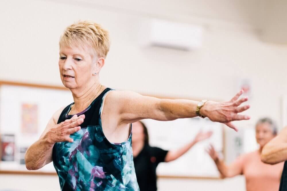 An elderly woman wearing a patterned tank top stretches her arms out in a fitness class. Other participants are in the background. - Home Instead