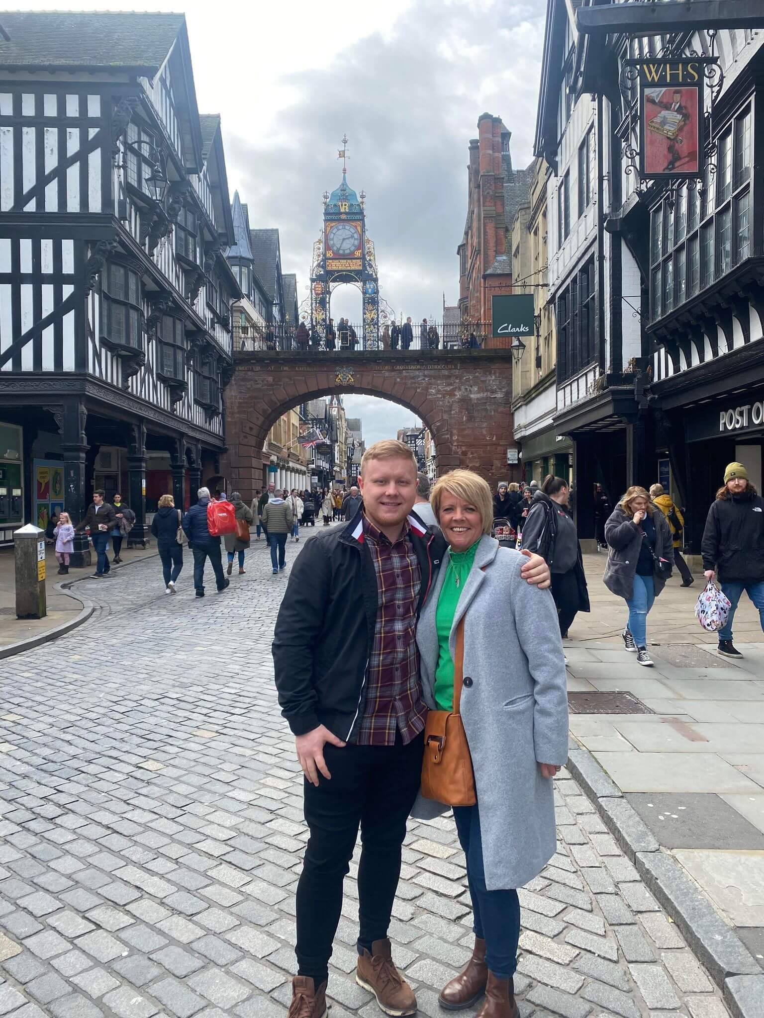 Two people standing on a cobblestone street in front of an ornate clock tower, surrounded by historical buildings. - Home Instead