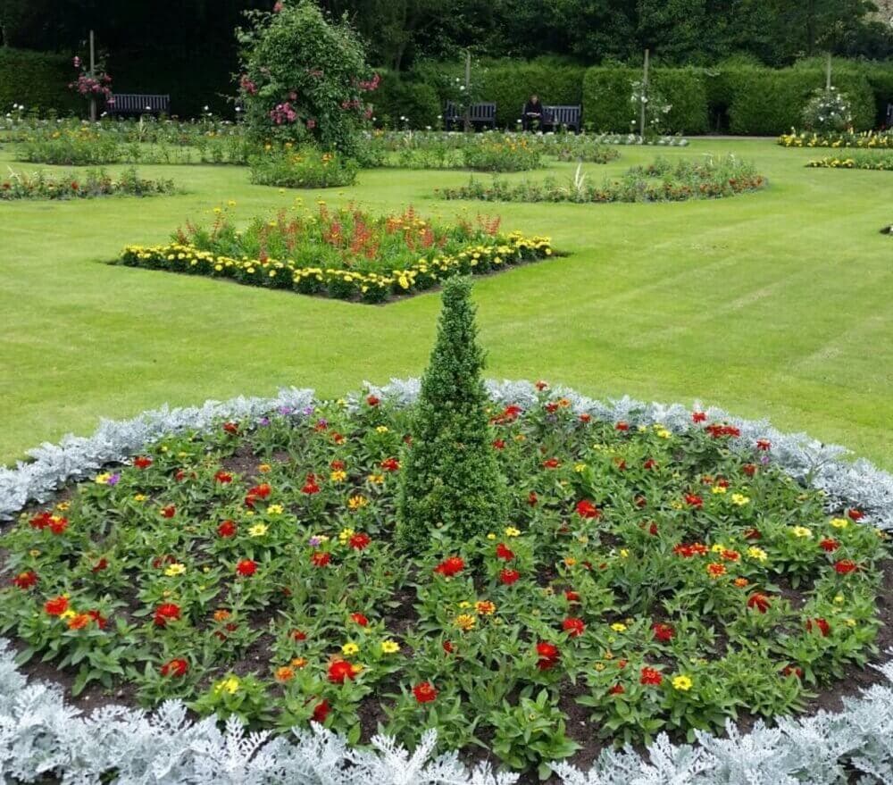 A formal garden with neatly trimmed lawns, flowerbeds in various shapes, and colorful flowers arranged in geometric patterns. - Home Instead