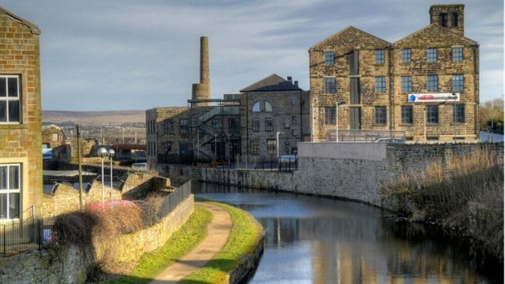 Industrial buildings and a tall chimney line a canal with a footpath on the banks, under a cloudy sky. - Home Instead