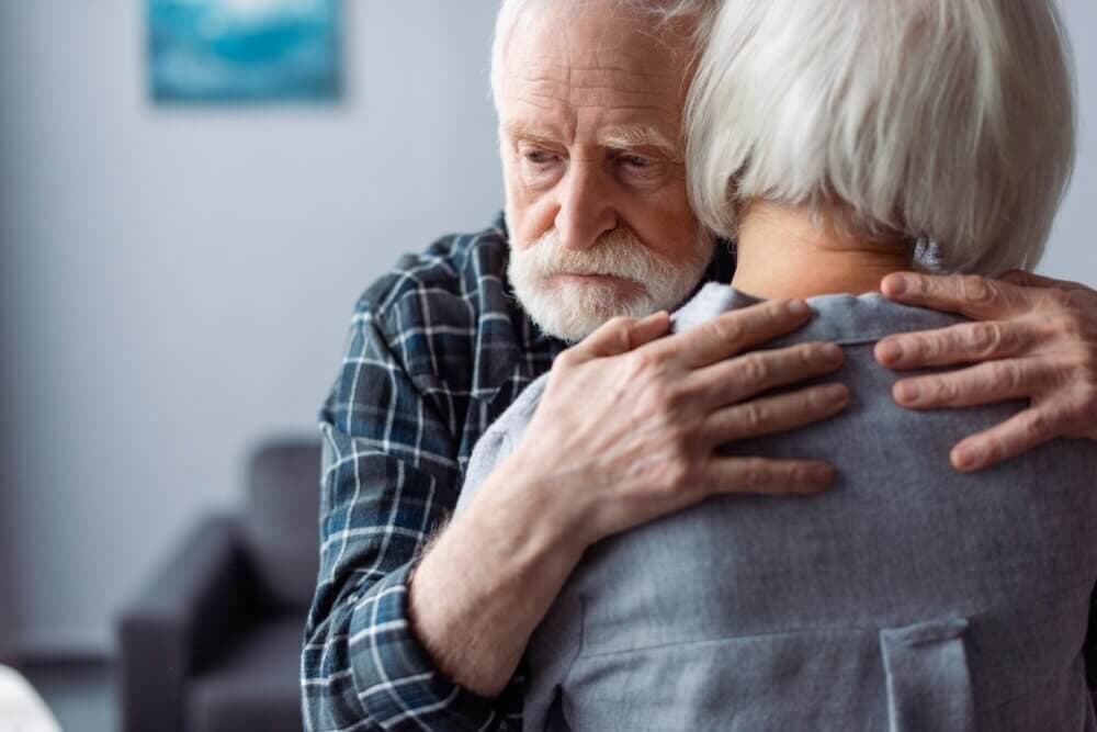 An elderly man with a somber expression embraces an elderly woman in a comforting hug in a living room. - Home Instead