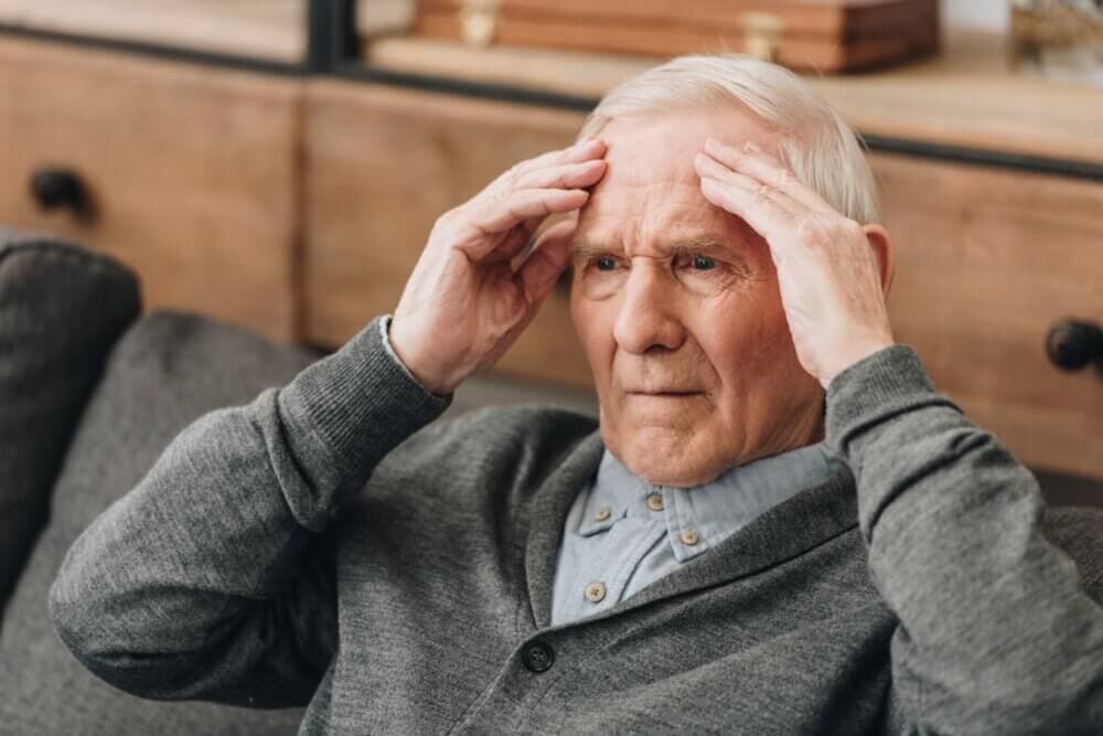 Elderly man sitting on a couch, holding his head with both hands and looking concerned, wearing a grey sweater. - Home Instead