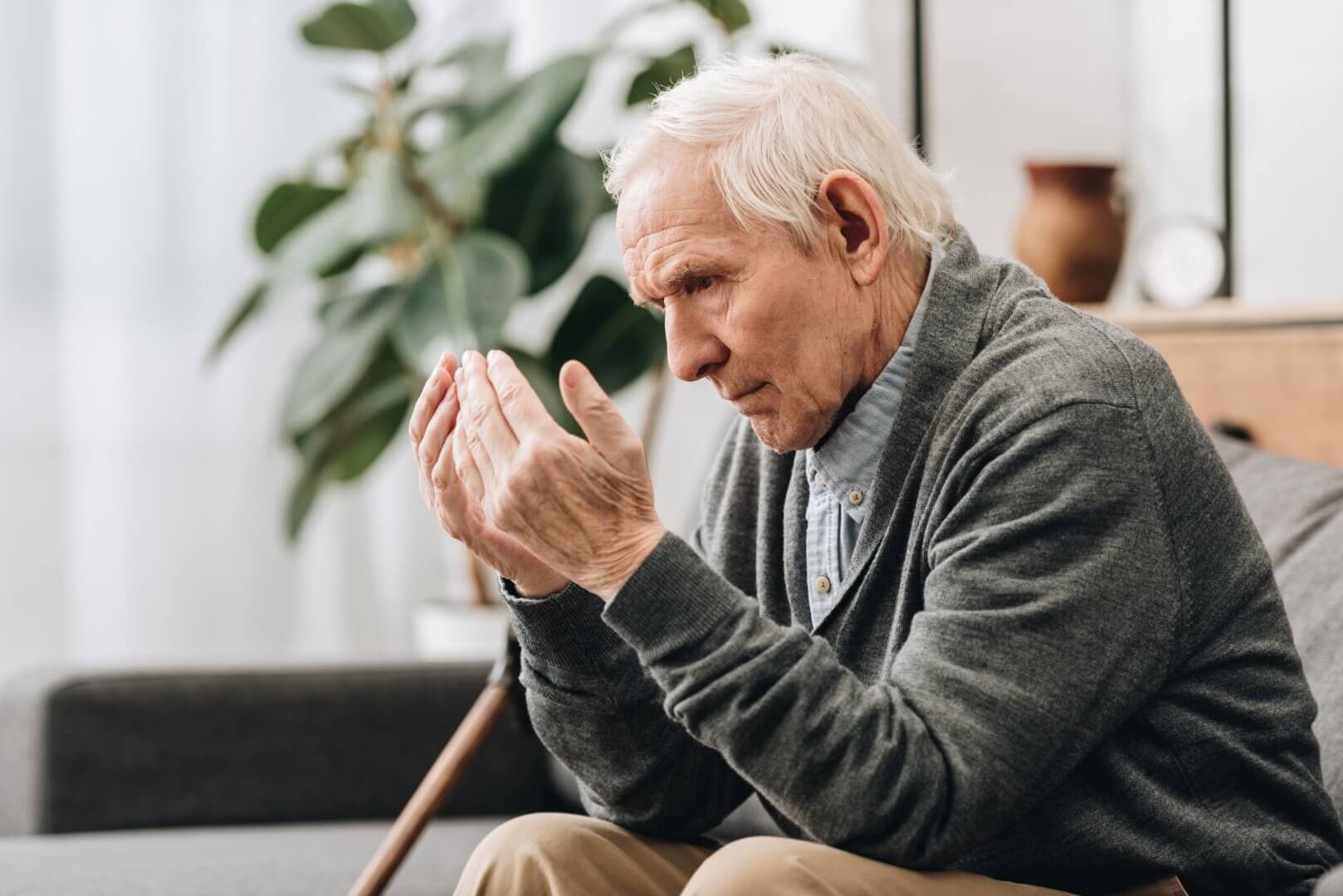 Elderly man sitting on a couch, looking at his hands with a cane by his side in a well-lit room. - Home Instead