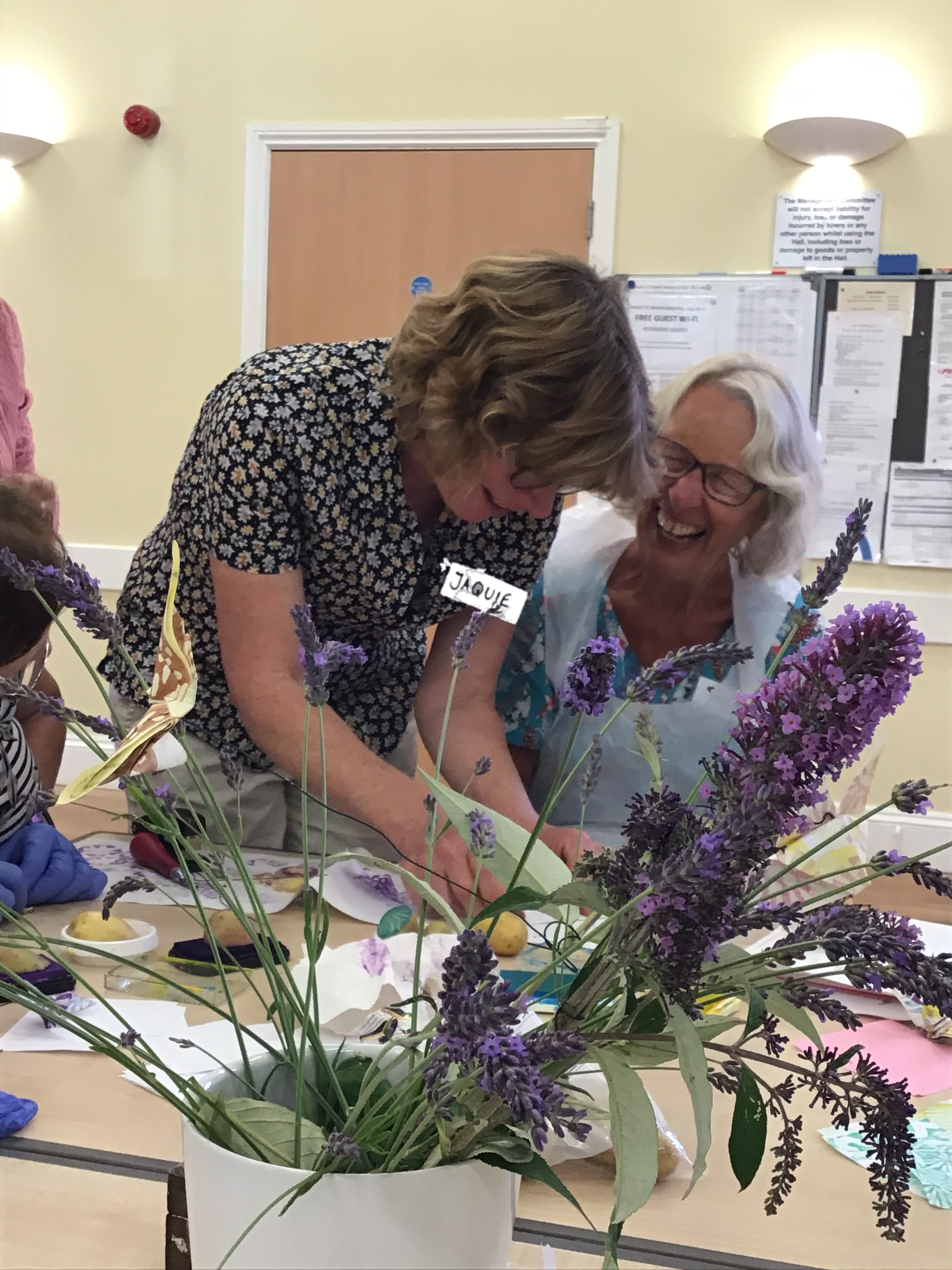 Two women laugh together while crafting at a table with lavender flowers in the foreground. - Home Instead