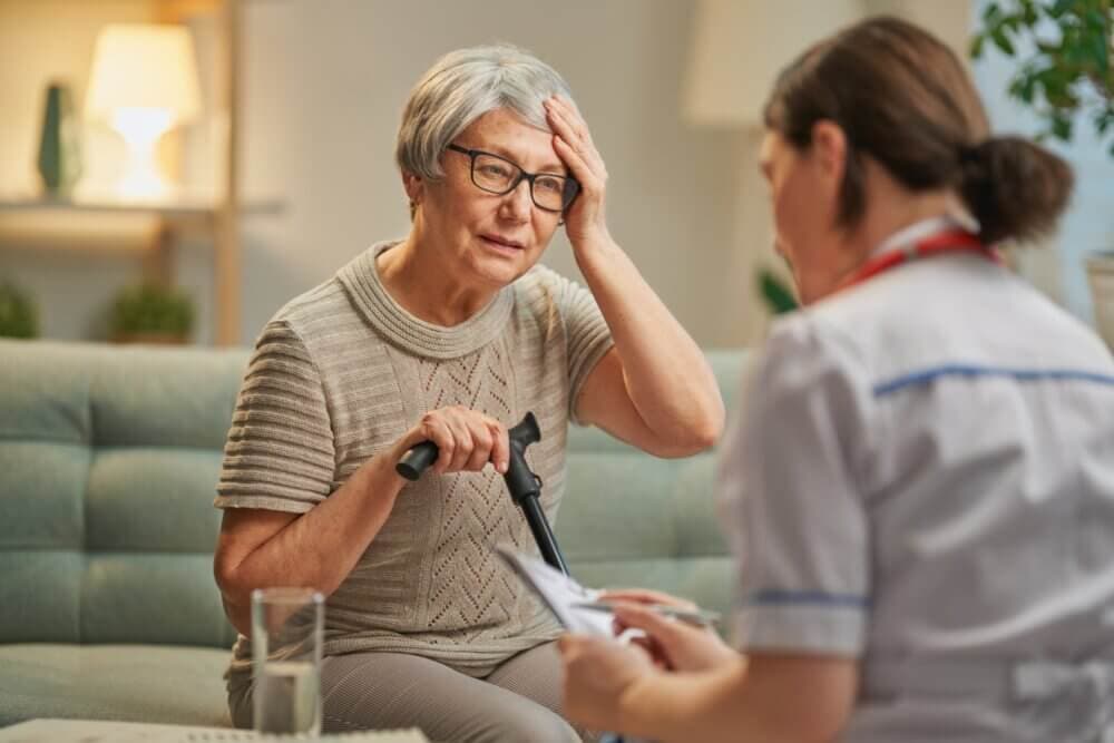 Elderly woman with a cane looks concerned, sitting on a couch, while a healthcare worker shows her paper documents. - Home Instead