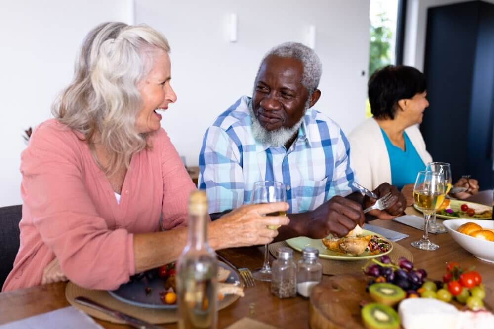 Three people smiling and eating together at a dining table with wine and food, including fruits and bread. - Home Instead