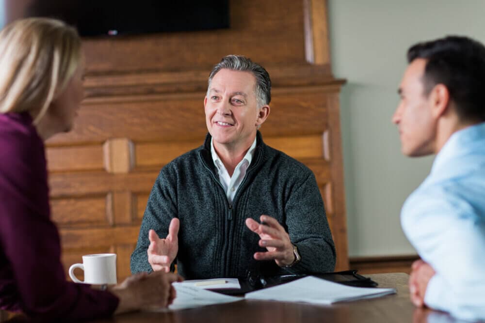Three people having a discussion around a table, one person gesturing with hands. A coffee mug is visible on the table. - Home Instead