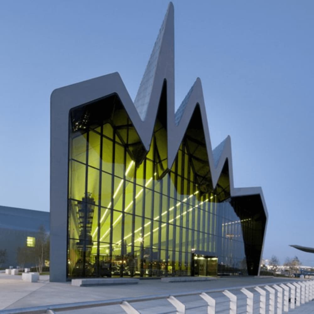 Modern building with a unique, jagged roof design and large glass facade illuminated by yellow lights at dusk. - Home Instead