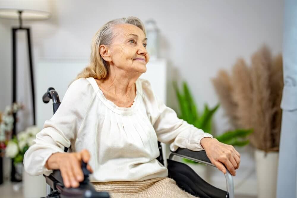 Elderly woman with light hair in a white blouse smiles while sitting in a wheelchair in a bright room with plants. - Home Instead