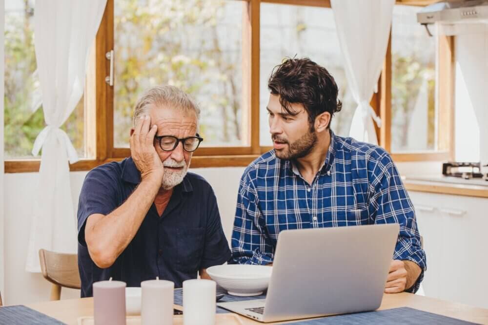 An older man sits with his hand on his face, and a younger man beside him points at a laptop screen in a sunlit kitchen. - Home Instead