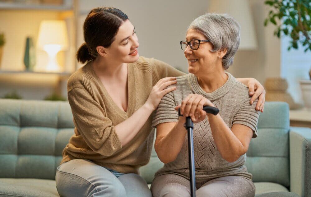 A younger woman comforts an elderly woman with a cane, both smiling and sitting on a light blue couch. - Home Instead