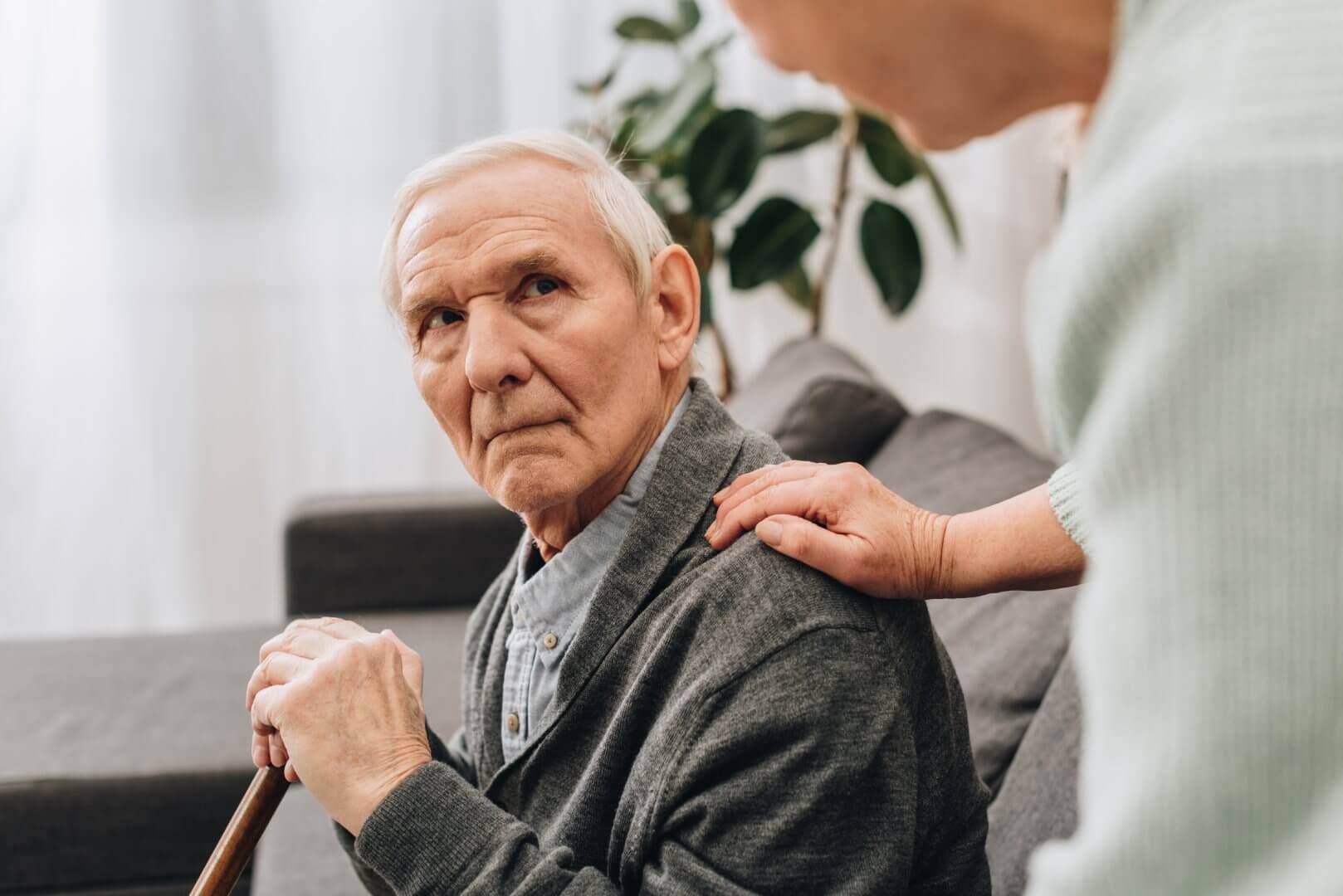 An elderly man sitting on a couch with a cane, looking up at someone who is comforting him with a hand on his shoulder. - Home Instead