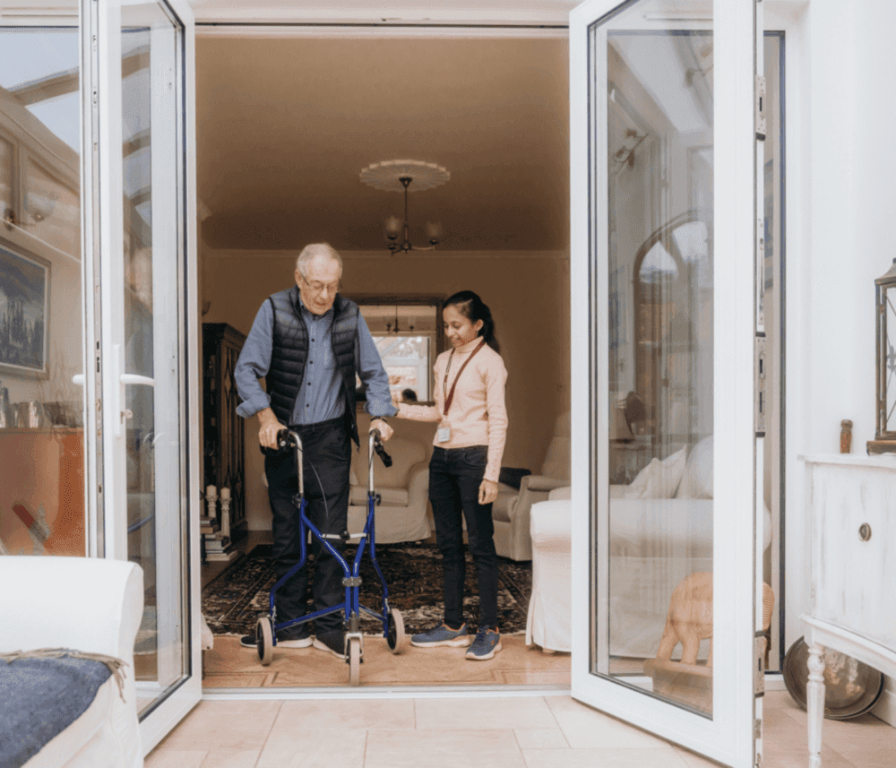An elderly man with a walker is assisted by a young woman as they step through a doorway into a cozy living room. - Home Instead