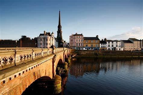 Scenic view of a historic bridge over a calm river, with quaint buildings and a tall spire in the background. - Home Instead